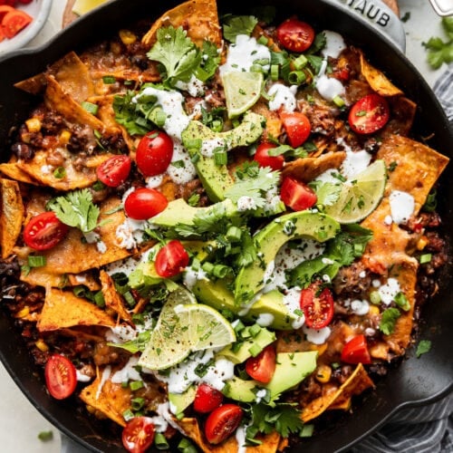 Overhead view skillet filled with beef enchiladas topped with avocado slices, sour cream and tomato slices.