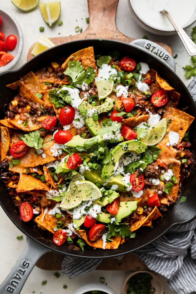 Overhead view cast iron skillet filled with beef enchiladas topped with avocado slices, sour cream and sliced tomatoes