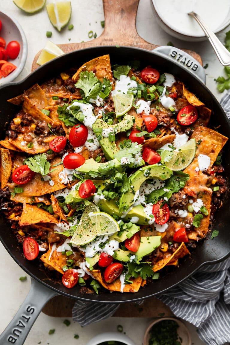 Overhead view skillet filled with beef enchiladas topped with avocado slices, sour cream and tomato slices.