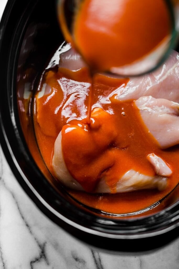 Buffalo sauce being poured over raw chicken in black slow cooker