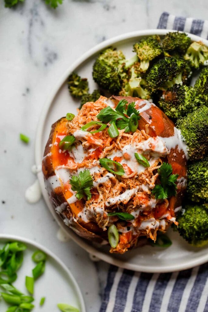 Overhead view slow cooker buffalo chicken shredded and stuffed into open baked sweet potato with side of roasted broccoli