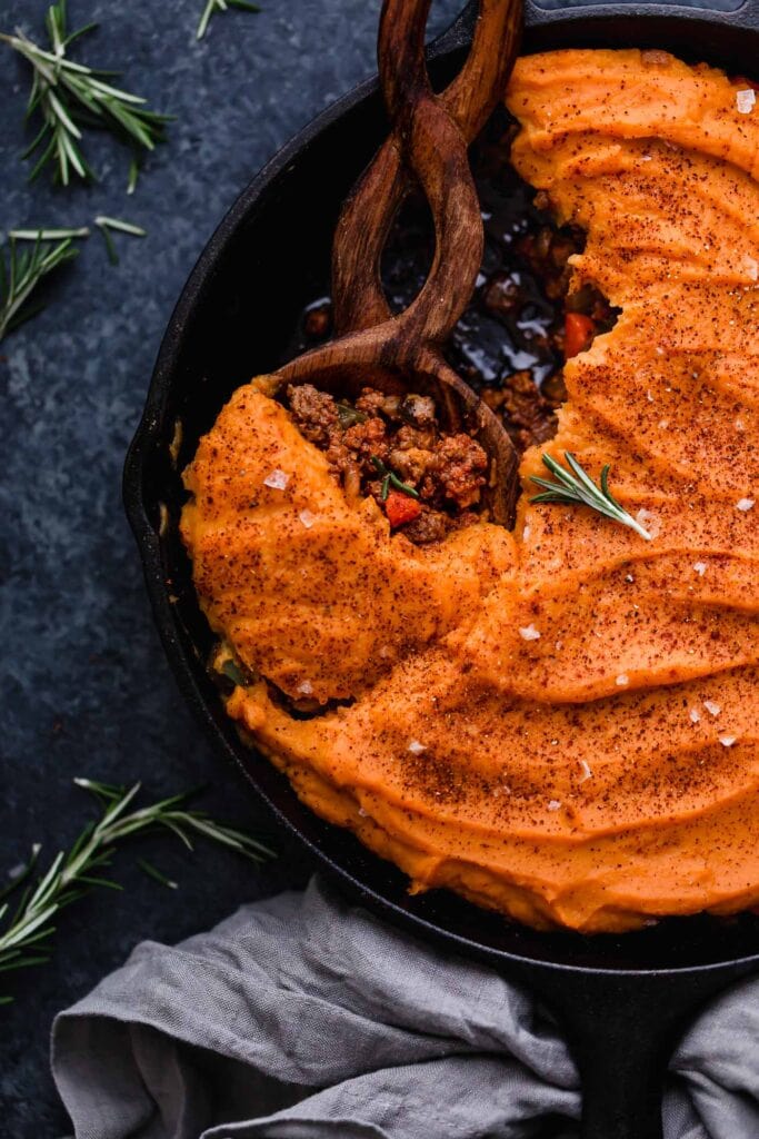 Overhead view cast iron skillet filled with shepherd's pie topped with mashed sweet potatoes, twisted handle wooden spoon scooping up serving