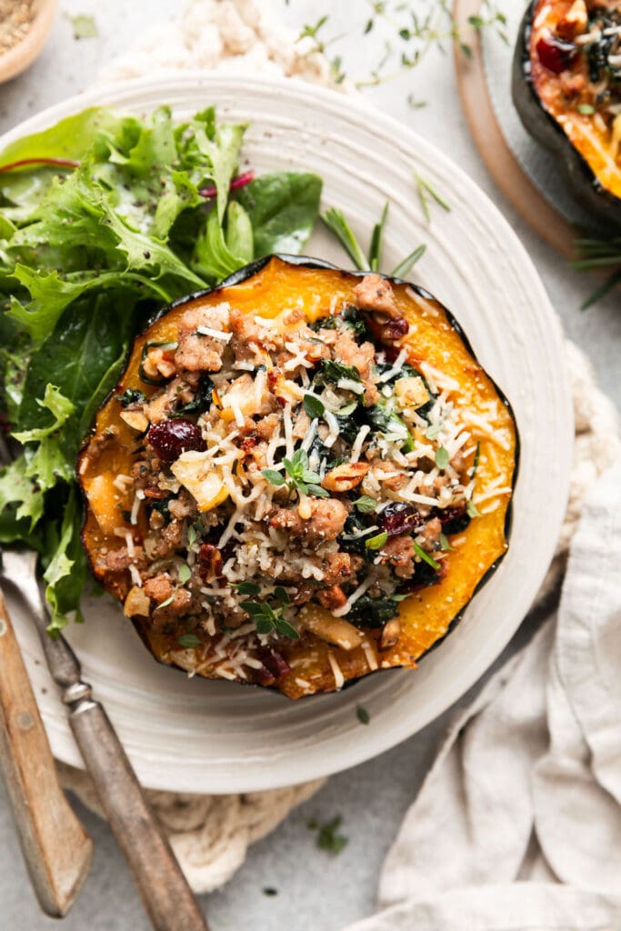 Overhead view sausage and spinach stuffed acorn squash half on white plate with side salad, topped with melted Parmesan cheese