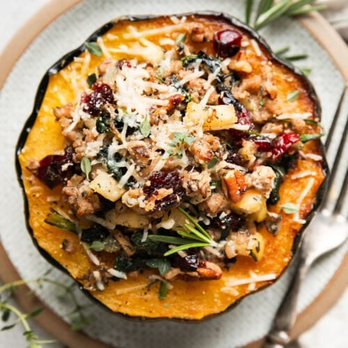 Overhead view sausage stuffed acorn squash half on plate, topped with Parmesan cheese, dried cranberries, pecans and fresh herbs
