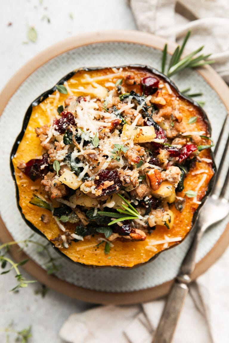 Overhead view sausage stuffed acorn squash half on plate, topped with Parmesan cheese, dried cranberries, pecans and fresh herbs