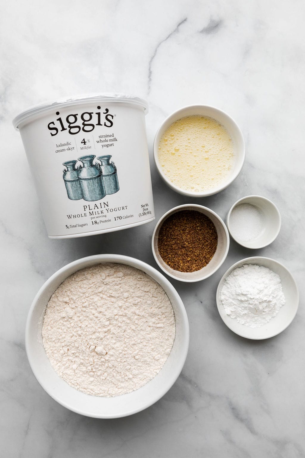 All of the ingredients to make Greek yogurt bagels shown from an overhead view. The ingredient include a container of Siggi's plain 4% yogurt, and ramekins of flour, baking powder, flax meal, salt, and egg whites.