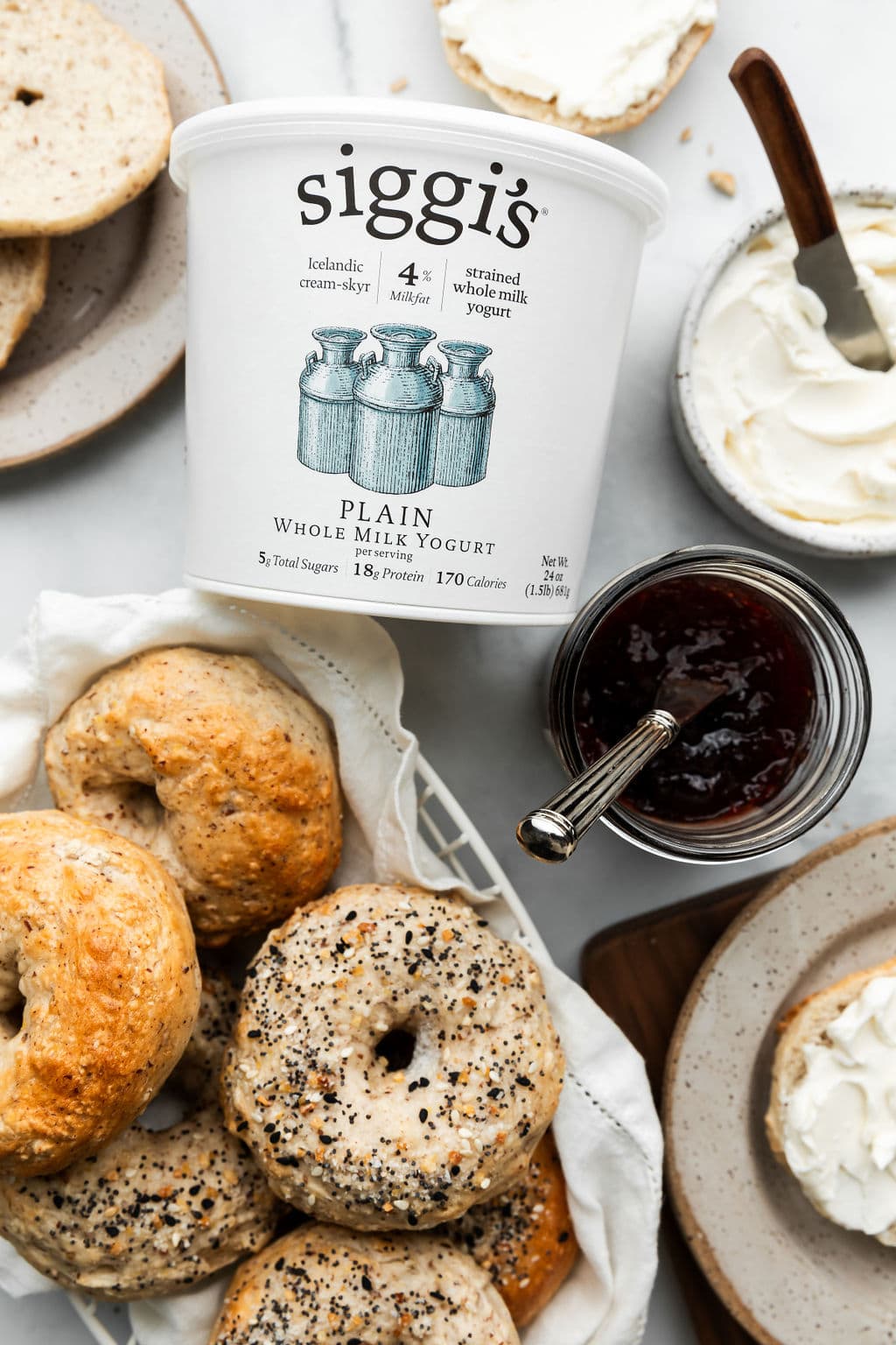 An overhead view of a counter with Greek yogurt bagels, a container of Siggi's plain 4% milkfat yogurt, jam and cream cheese both with knives in the containers.