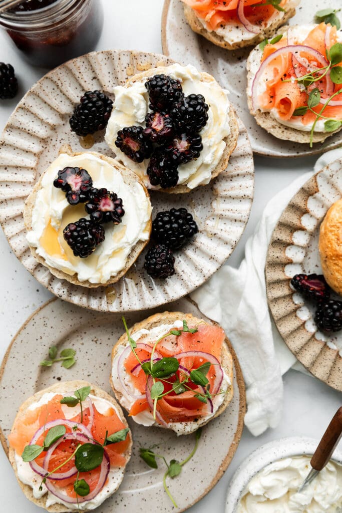 A pair of Greek yogurt bagels each on separate plates. One is topped with cream cheese, fresh blackberries and a drizzle of honey, while the other is topped with cream cheese, smoked salmon lox, thinly sliced pickled red onions and watercress. 