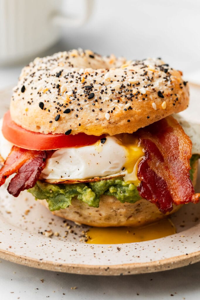 An everything Greek yogurt bagel breakfast sandwich on a plate. The sandwich is made of avocado, bacon, tomato, and a fried egg with the yolk escaping to the plate. 