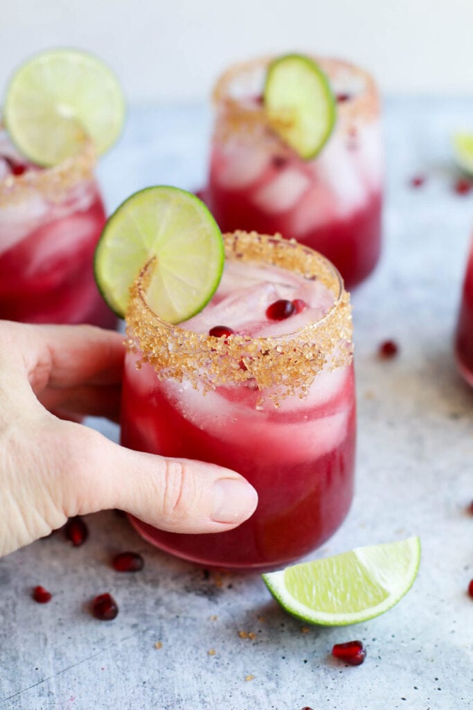 A hand holding a glass of pomegranate margarita, complete with a sugared rim and lime wheel. 