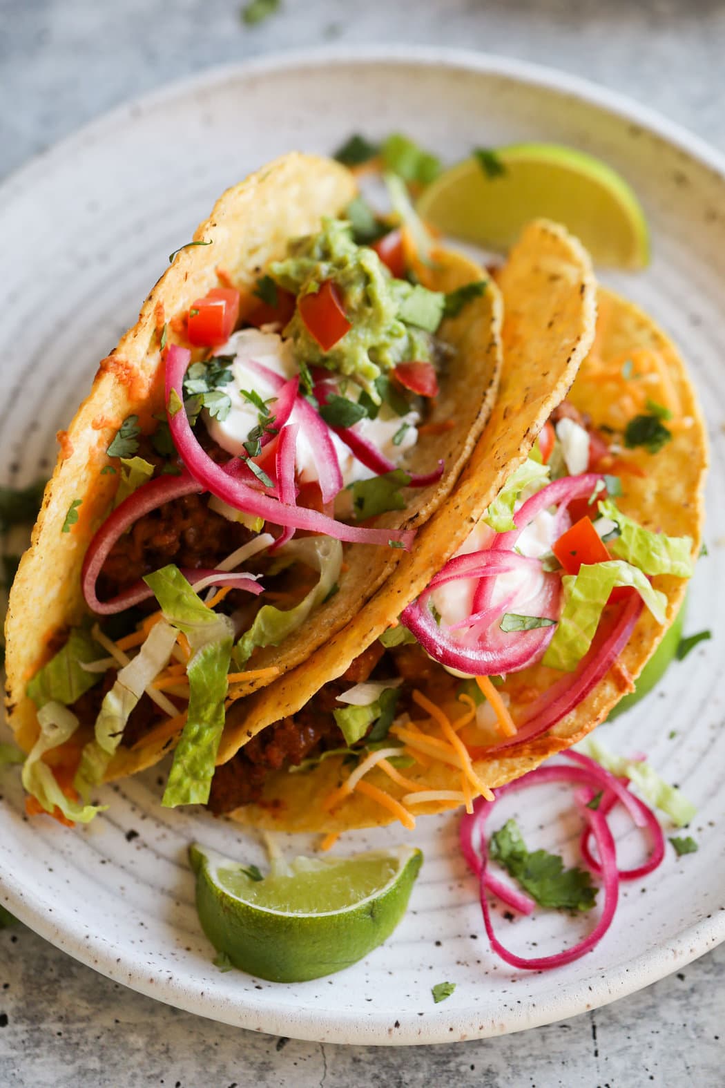 Close up view of two fully prepared baked tacos on a white plate topped with fresh guacamole, pickled red onions, and sour cream