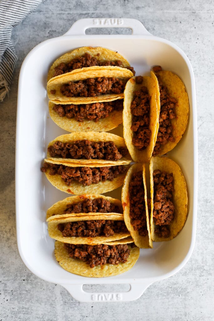 Overhead view of yellow taco shells filled with ground taco meat in a white baking dish. 