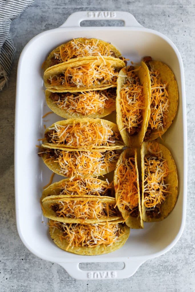 Overhead view of yellow taco shells filled with taco meat and shredded cheese arranged in a white baking dish. 