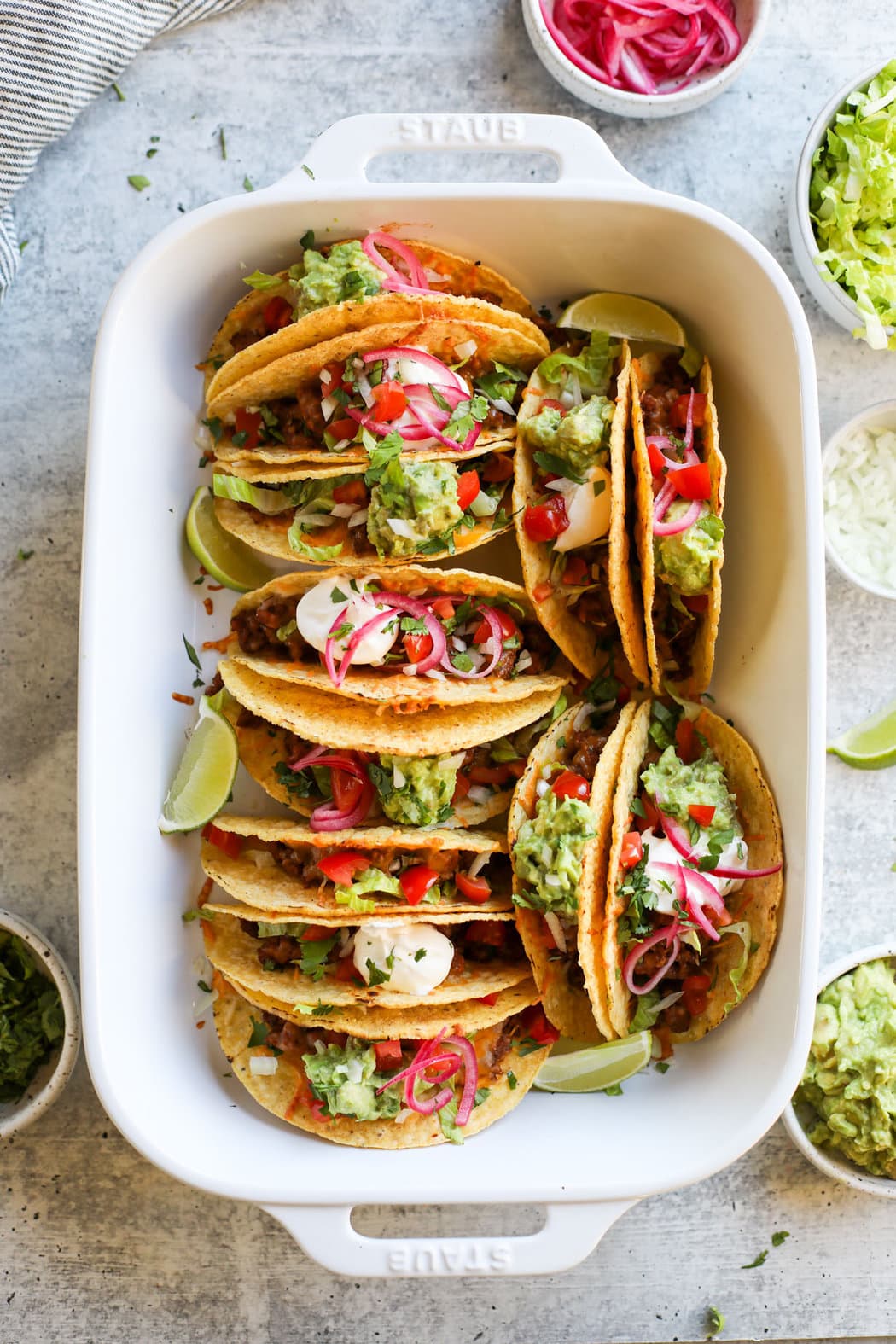 Overhead view of a white baking dish filled with baked tacos and topped with fresh guacamole and garnished with limes. 