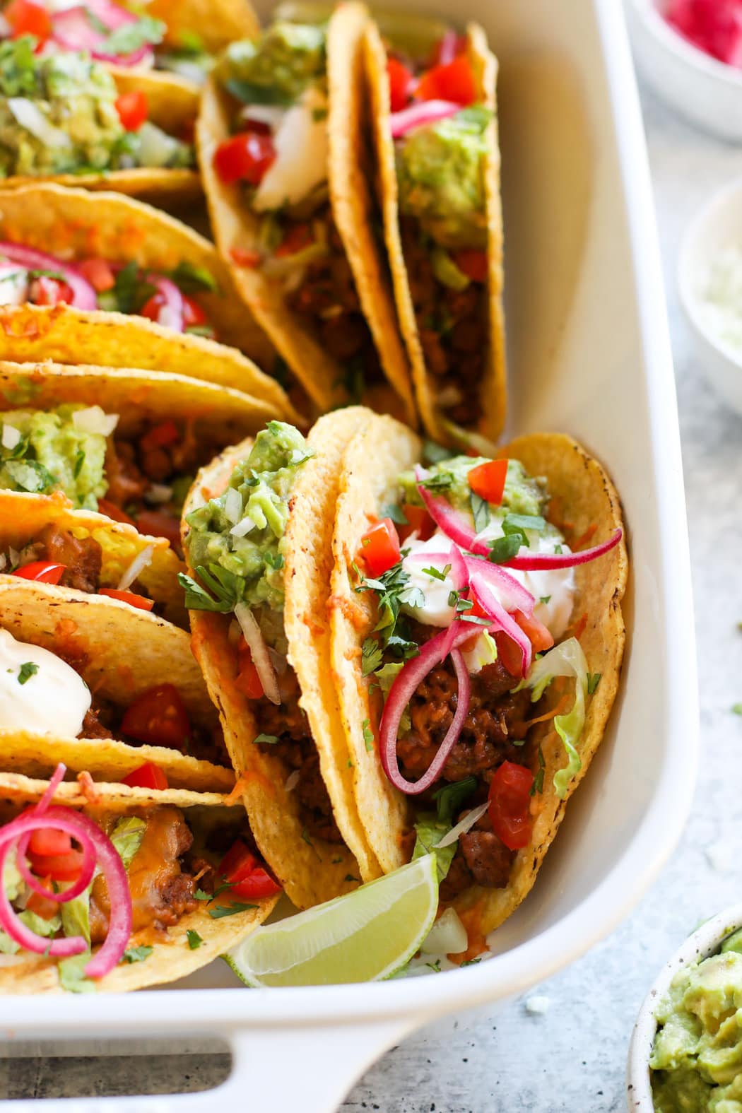 Close up view of white baking dish filled with baked tacos garnished with fresh toppings and bright red sliced onions. 