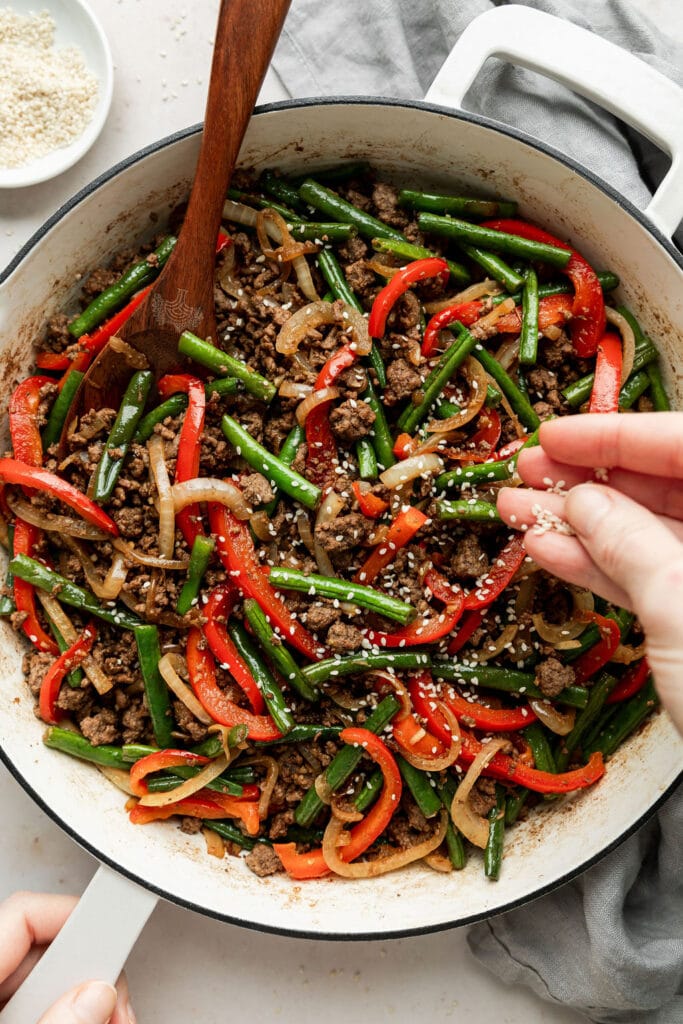 Overhead view of white skillet containing Ground Beef Stir Fry and a hand sprinkling sesame seeds on top. 