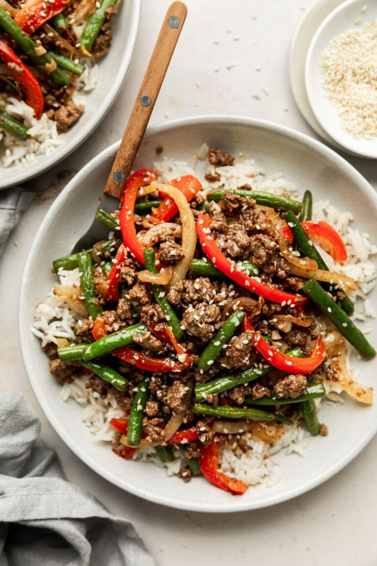 Overhead view of a white plate containing ground beef stir fry on top of white rice and topped with sesame seeds. 