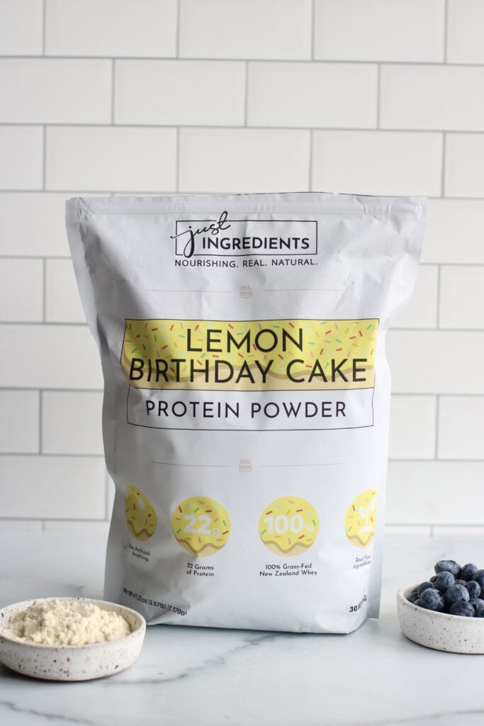 A bag of Just Ingredients Lemon Birthday Cake protein powder on a marble counter in front of a white tiled backsplash. 