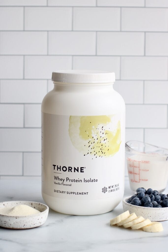 A large container of Thorne Whey Protein Isolate (vanilla flavor) on a counter with a measuring cup of milk, some blueberries, and sliced bananas in front of a white tiled backsplash. 
