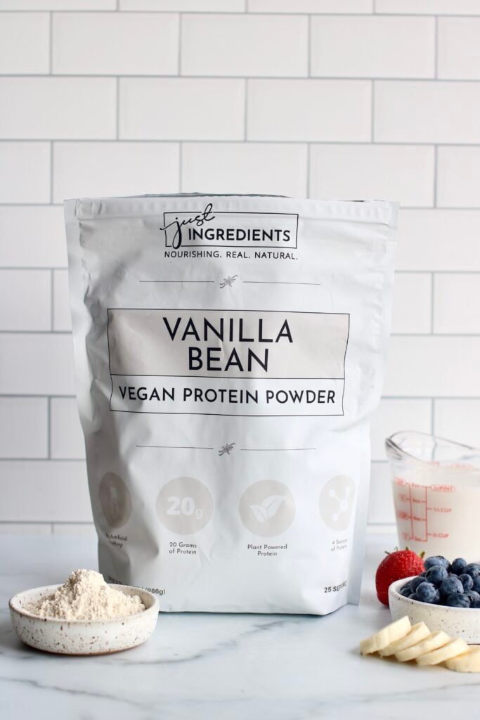 A bag of Just Ingredients Vegan Vanilla Bean protein powder on a white marble surface with milk, berries, and banana slices in front of a white tiled backsplash. 