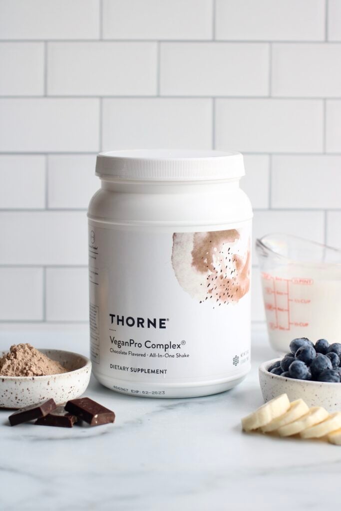 A carton of Thorne VeganPro Complete protein powder on a white marble surface with milk, berries, banana slices, cocoa powder, and chocolate in front of a white tiled backsplash. 