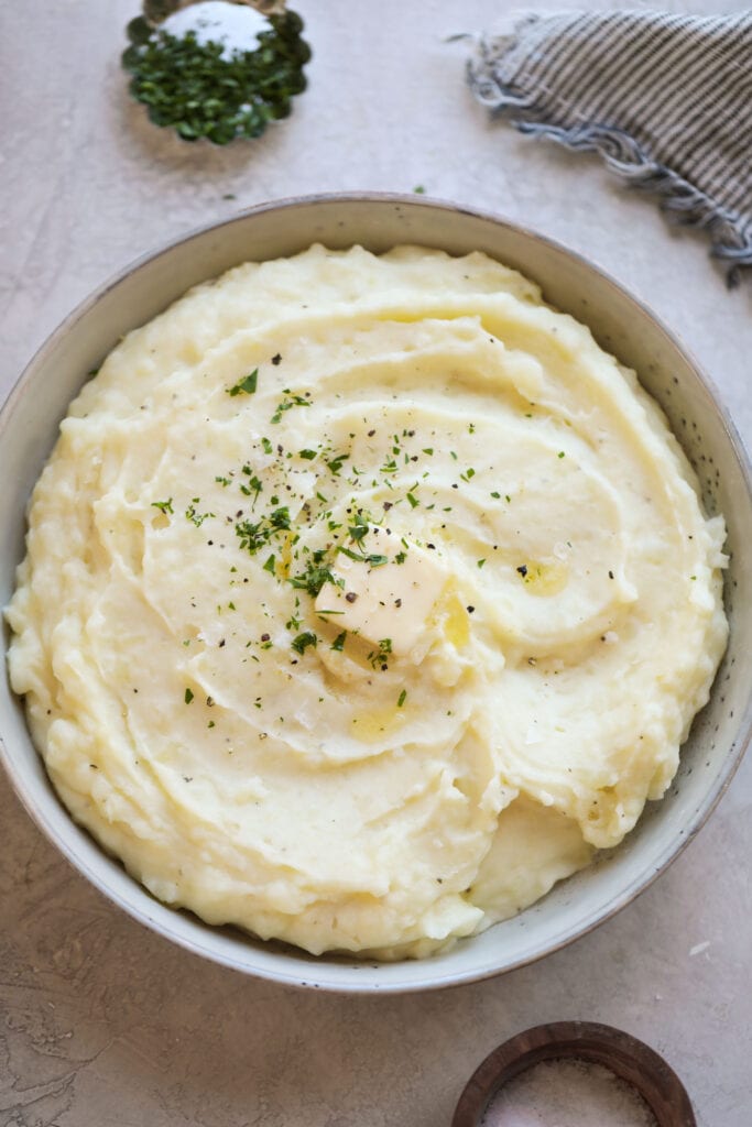 Overhead view of mashed potatoes in a white bowl topped with melting butter and fresh herbs. 
