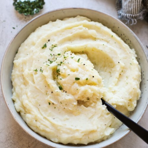 White bowl of Mashed Potatoes topped with butter and fresh herbs.