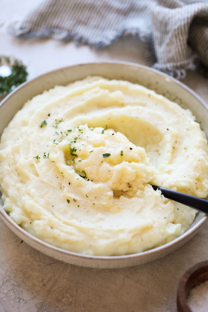 Overhead view of a white bowl filled with mashed potatoes being scooped with a large spoon. 