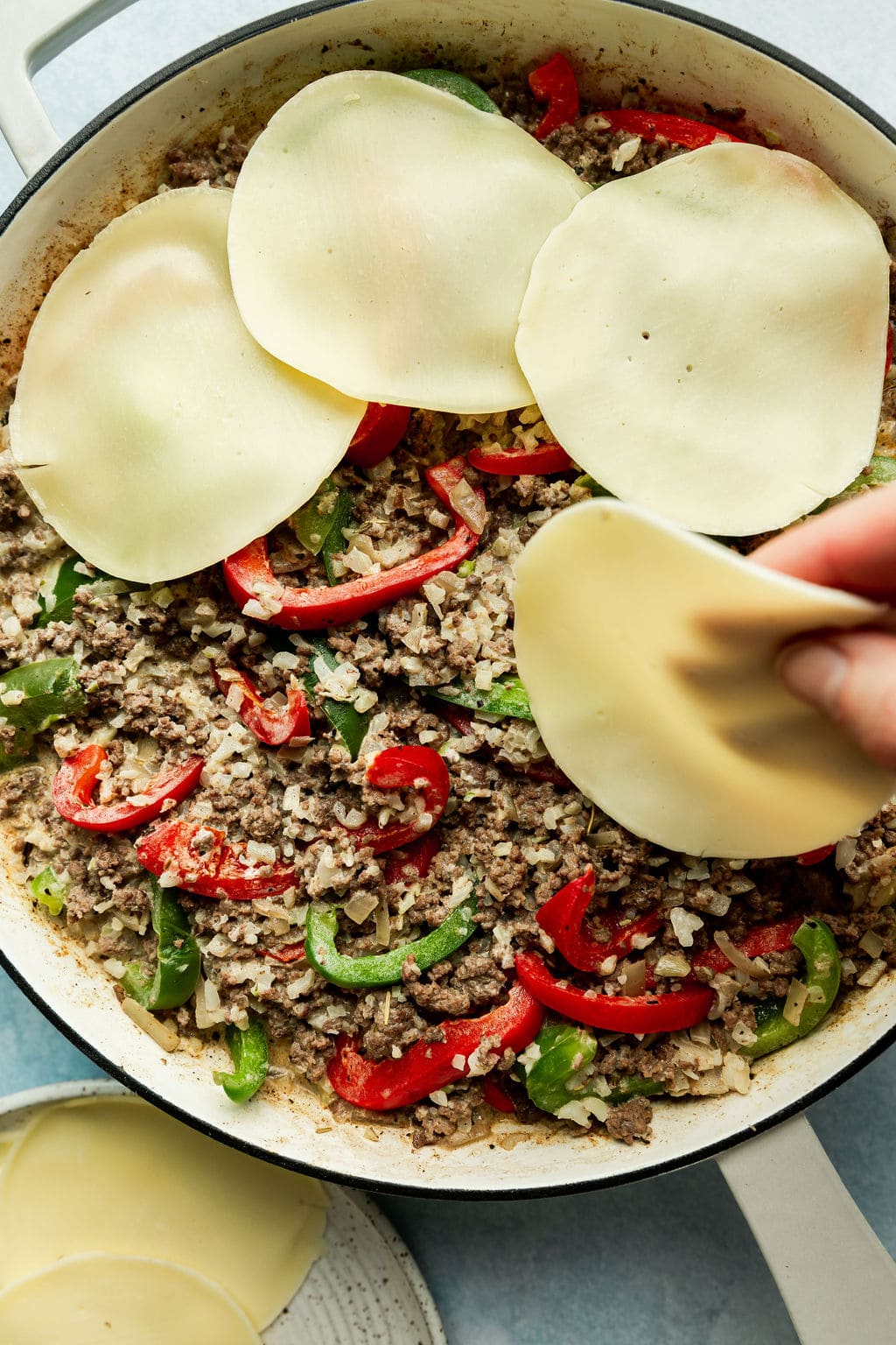 Overhead view of white Skillet containing half cooked Low Carb Philly Cheesesteak Casserole being topped with large circular pieces of white cheese ready to melt. 
