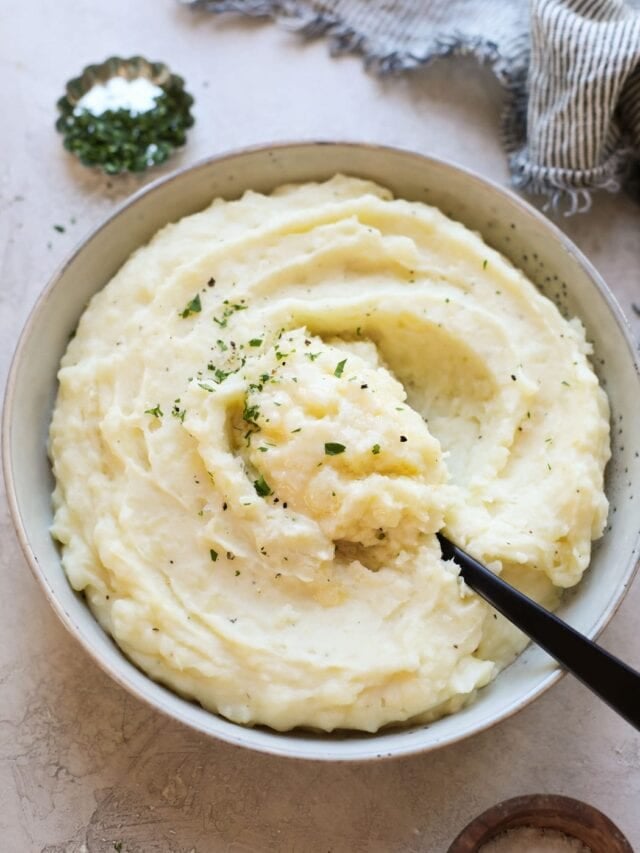 White bowl of Mashed Potatoes topped with butter and fresh herbs.