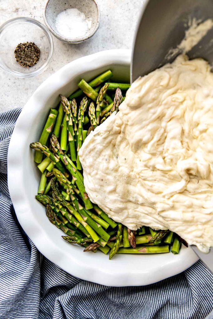 Pouring sour cream mixture from a skillet over fresh asparagus in a dish. 