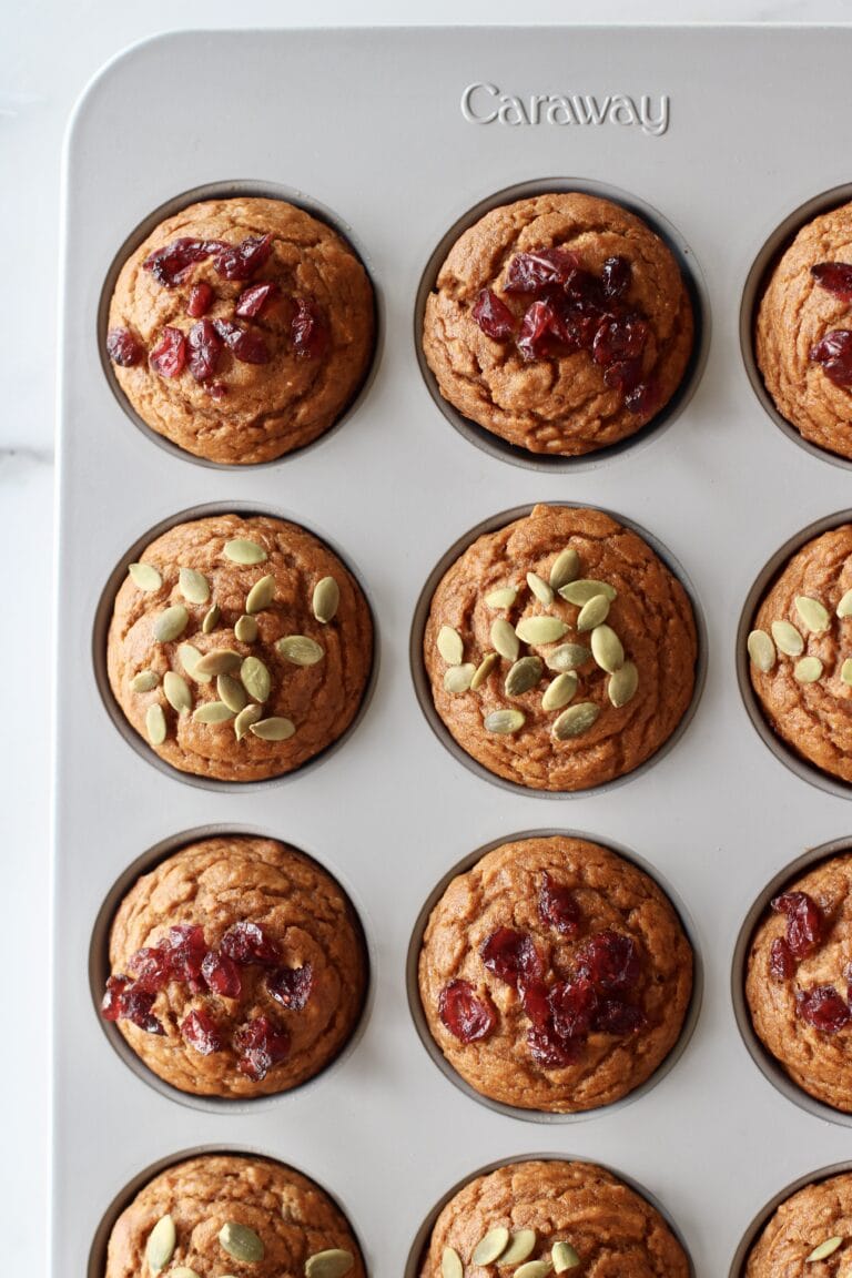 Overhead view of a freshly baked pan of pumpkin muffins topped with a variety of pumpkin seeds and dried cranberries. 