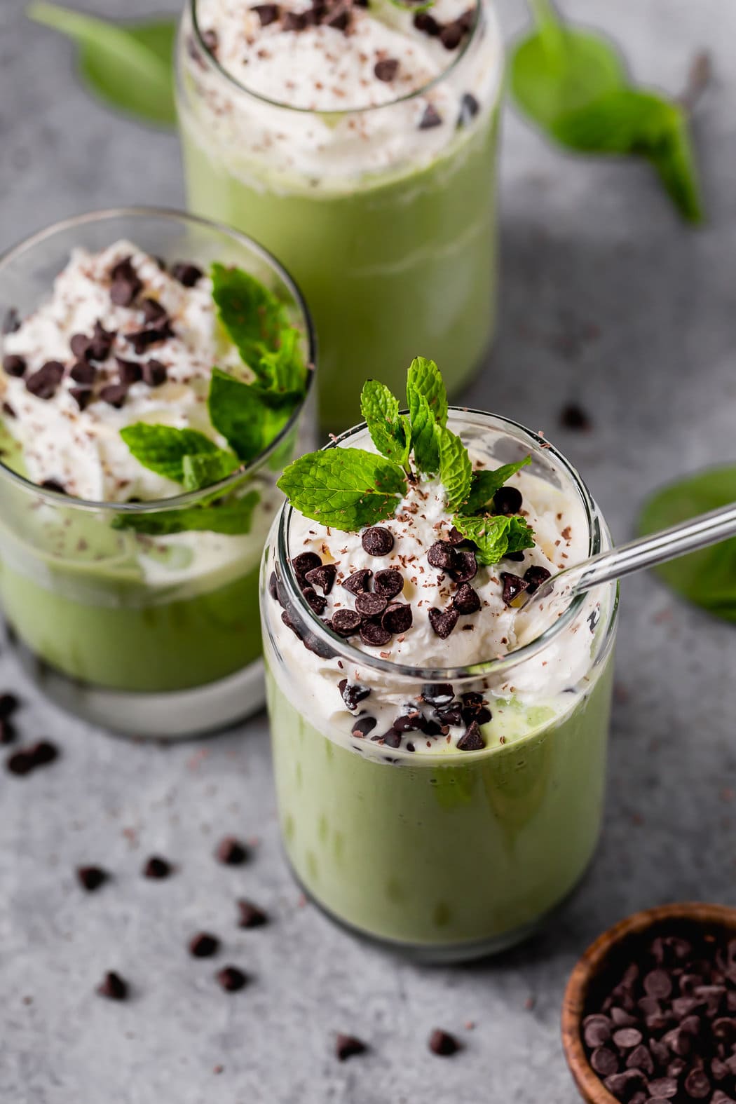 Close up view of vibrant green shamrock shakes in glasses topped with whipped cream, chocolate chips, and fresh mint. 