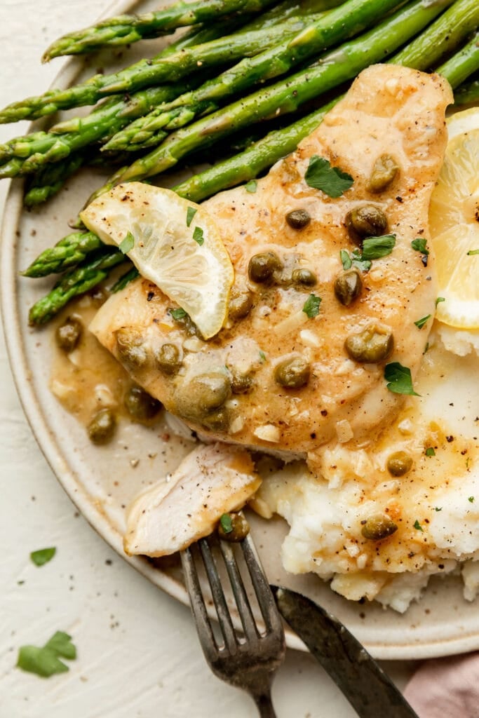 A white plate with a portion of chicken piccata served over mashed potatoes and sautéed asparagus. 
