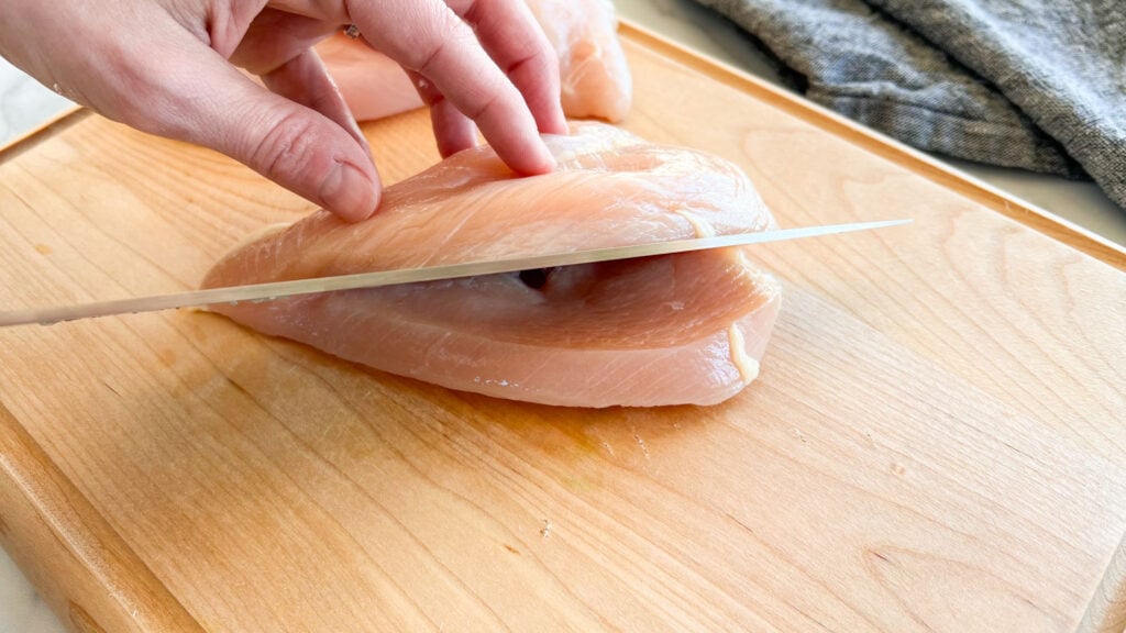 A boneless skinless chicken breast on a cutting board with a hand using a large knife to cut it in half horizontally as if butterflying the chicken breast.