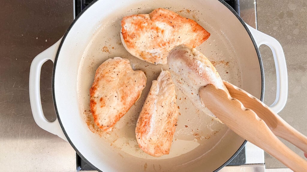 A white skillet with 4 chicken breasts browning in it. One of the breasts is held in the air with a pair of tongs while it is being flipped over.