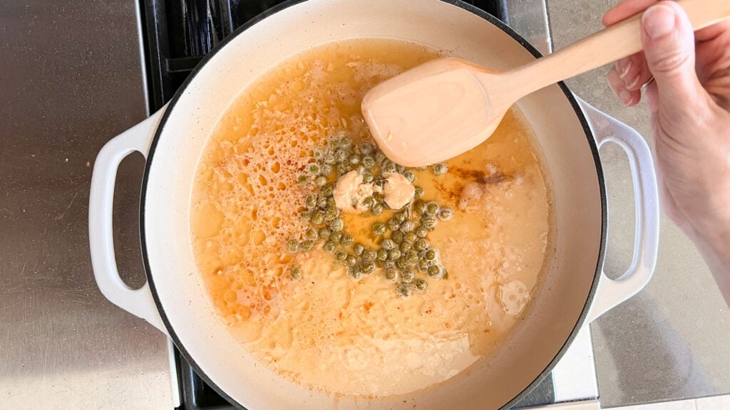 A white skillet on the stovetop with broth, mustard, and capers. 