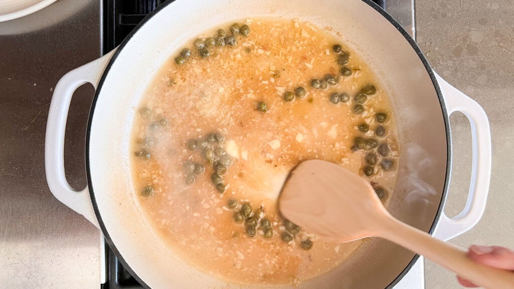 A wooden spoon stirring the lemon-caper sauce while it thickens slightly. 