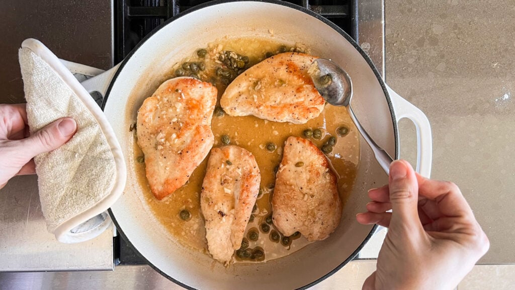 A hand spooning the lemon-caper sauce over the cooked chicken breasts in a white skillet. 