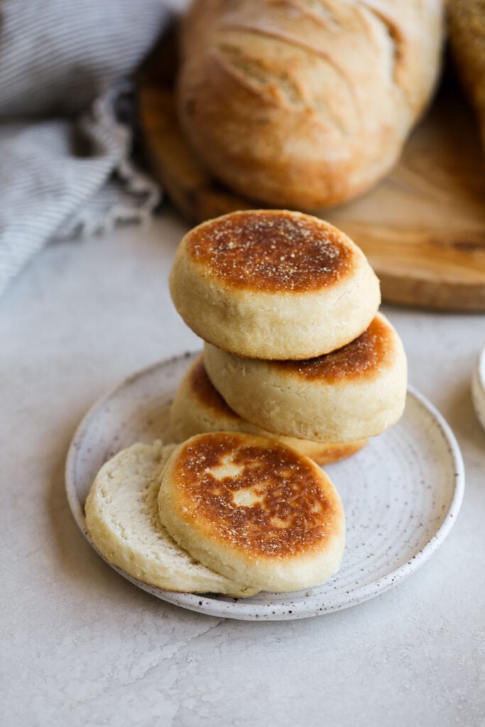 White plate of three stacked English muffins with a split English muffin in the foreground. 