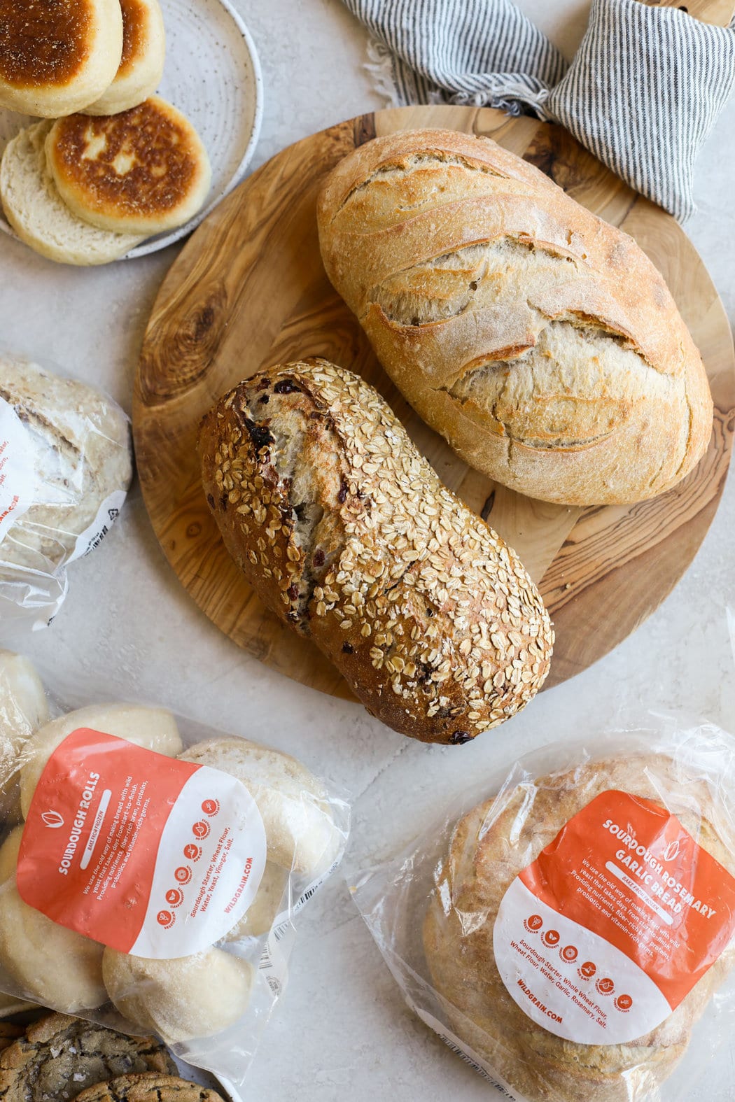 Overhead view of 2 loaves of sourdough bread on a wooden cutting board, surrounded by different sourdough products. 