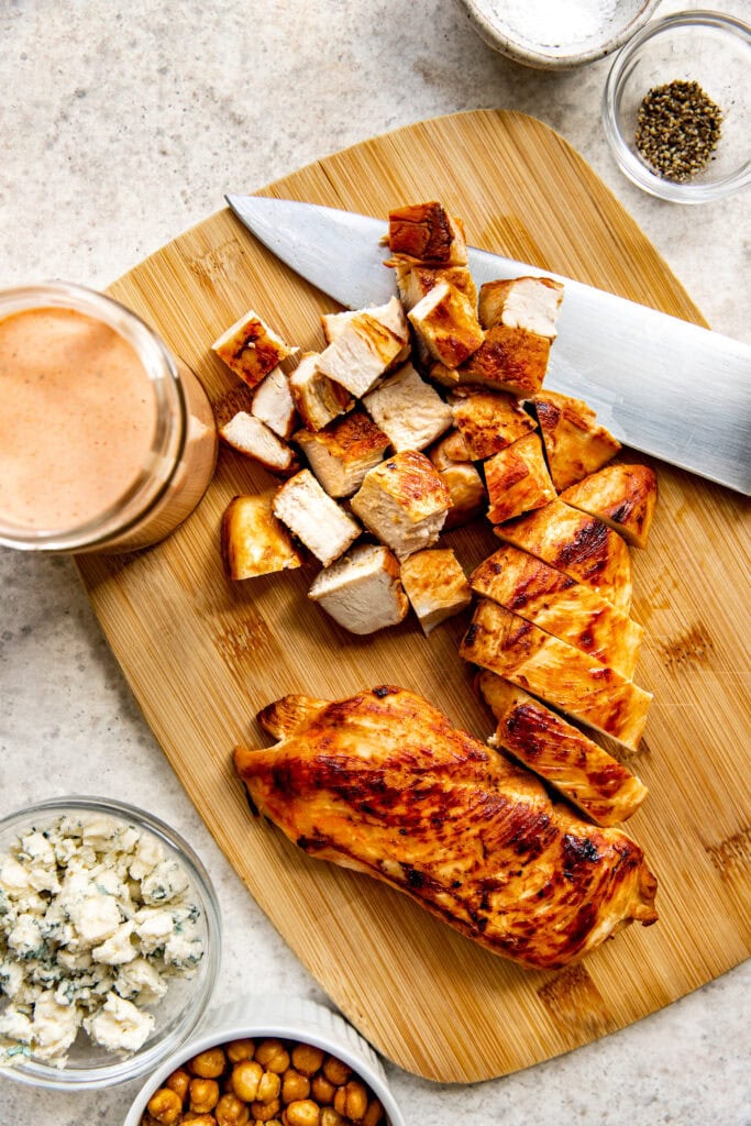Overhead view of grilled chicken being diced with a large knife on a wood cutting board. 