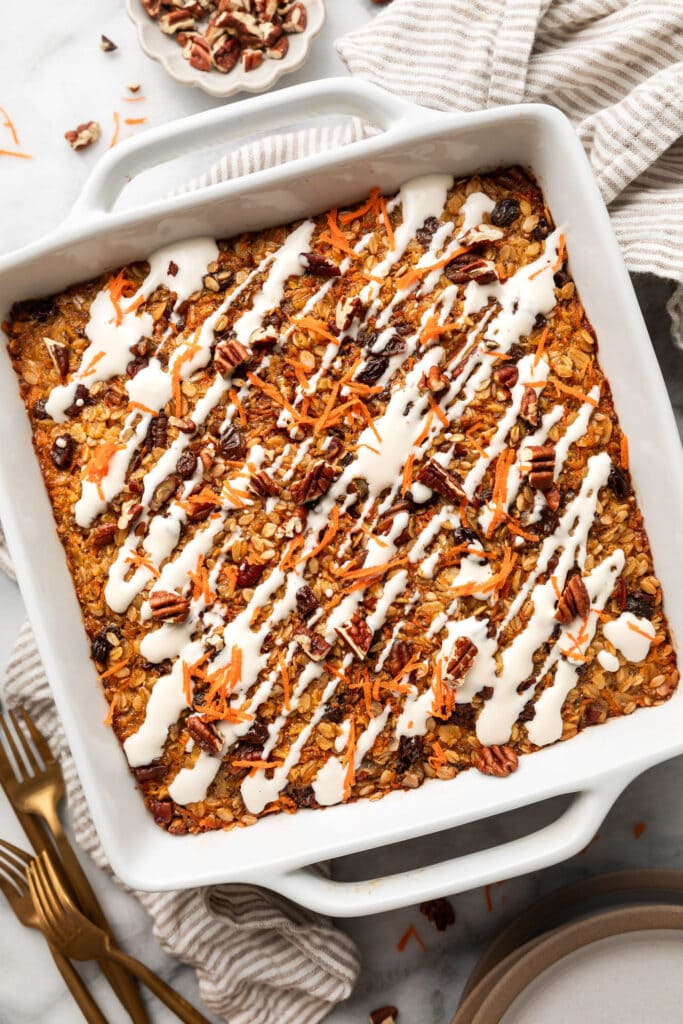 Overhead view of a white 9x9 baking dish with freshly baked carrot cake baked oatmeal topped with cream cheese drizzle, chopped pecans, and shredded carrots. 