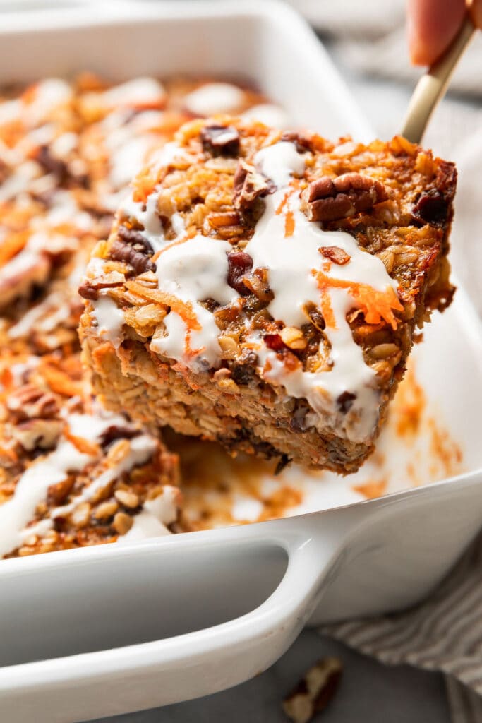 Close up view of a hand scooping a large piece of carrot cake baked oatmeal topped with cream cheese drizzle, pecans, and shredded carrots from a white baking dish.