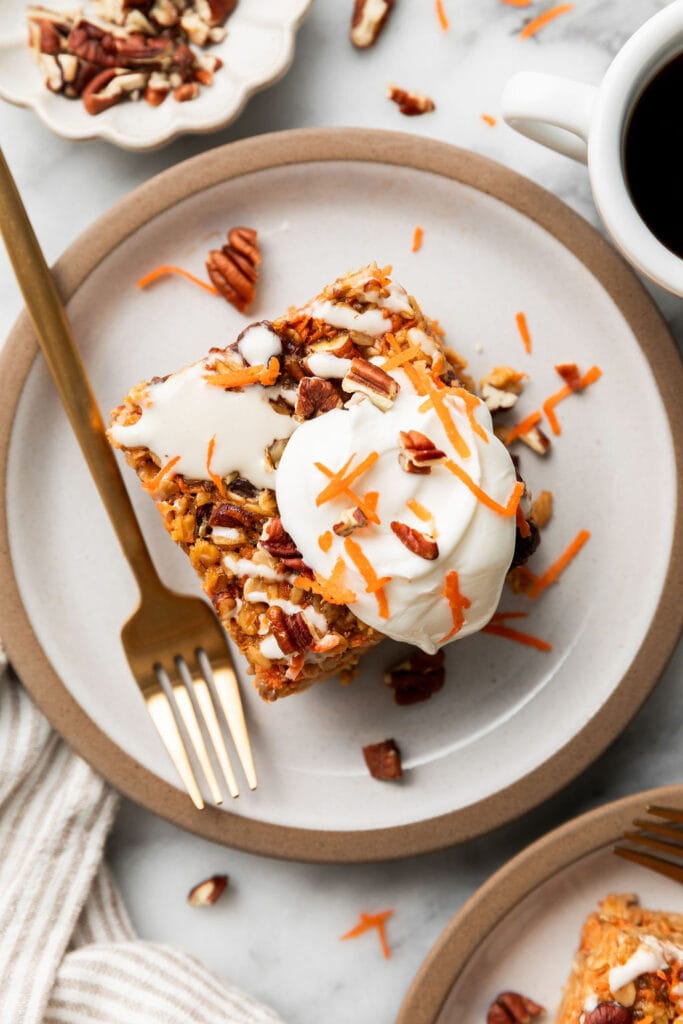 Overhead view of piece of carrot cake baked oatmeal on a white plate topped with a cream cheese drizzle, pecans, and shredded carrots. 