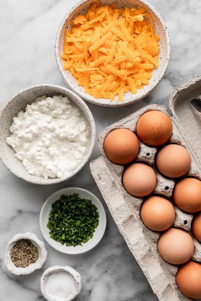 Overhead view of ingredients for egg bites including eggs, shredded cheddar, cottage cheese, chives, salt, and pepper. 