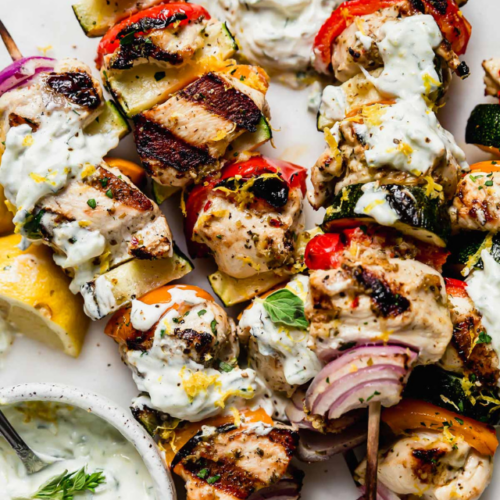 Overhead view Greek chicken and veggie kebabs on white platter with side of tzatziki sauce.