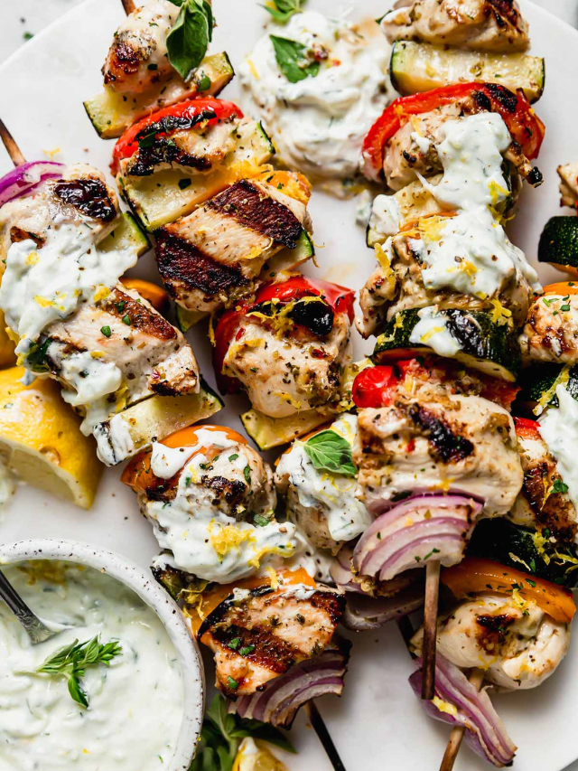 Overhead view Greek chicken and veggie kebabs on white platter with side of tzatziki sauce.