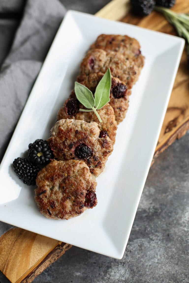 Blackberry breakfast sausages lined up on a white plate garnished with sage. 