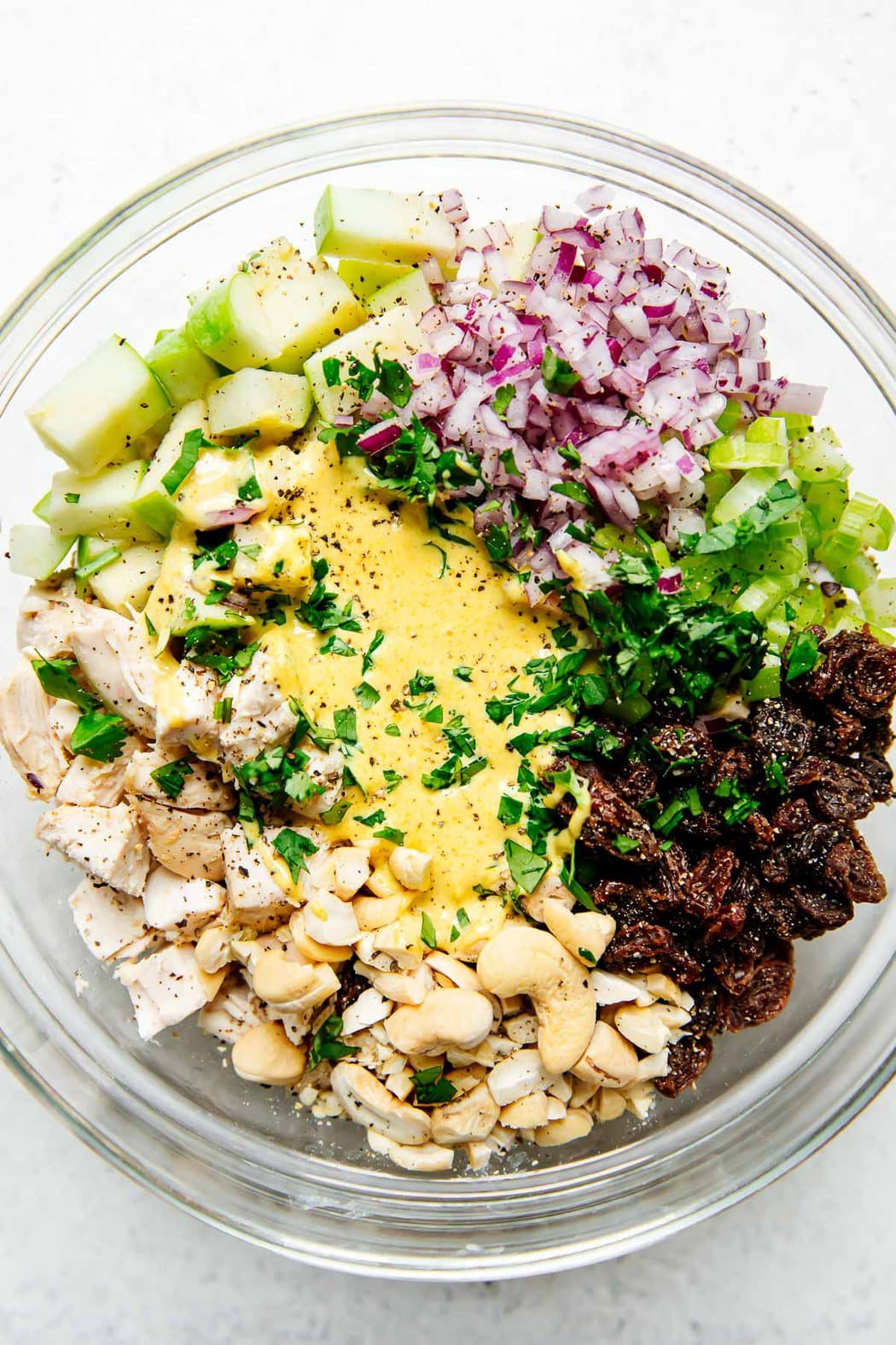 All ingredients for curry chicken salad in a bowl ready to be mixed together - cooked chicken, dressing, apples, red onion, celery, raisins and cashews.